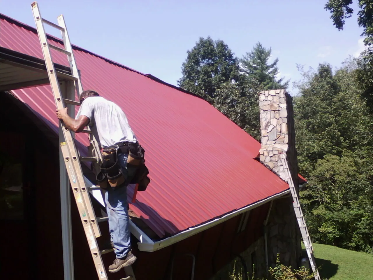 Roofer on a ladder beside a red metal roof and stone chimney — Soffit & Fascia Repair by Roofing Brothers in Green Valley